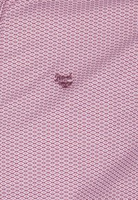 Rood en wit gestreepte stof met een diamanten patroon, waarop een klein logo in donkere draad is geborduurd nabij het midden.