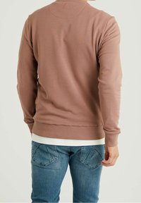 Sudadera marrón con un cuello y puños acanalados, que presenta una superficie texturizada y un pequeño detalle bordado en la parte posterior. Llevada sobre jeans azules.
