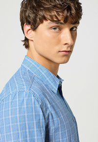 Camisa a cuadros azul y blanca con cuello abotonado y mangas cortas; el tejido parece ligero y con una textura suave.