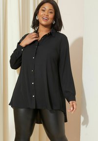 Schwarzes Langarm-Hemd aus weichem Stoff, mit silbernen Knöpfen, klassischem Kragen und abgerundetem Saum. Kombiniert mit schwarzen Leggings.