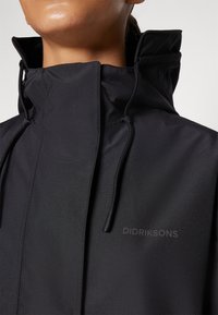 Schwarze wasserdichte Jacke mit hohem Kragen, verstellbaren Kordeln und dezentem Logo. Glatte Textur, minimalistisches Design und verdeckte Verschlüsse.