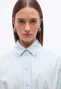 Νεαρή γυναίκα με ίσια καστανά μαλλιά, που φορά ασημένια σκουλαρίκια κρίκους και άσπρο πουκάμισο με λεπτές κατακόρυφες πράσινες και μπλε ρίγες.