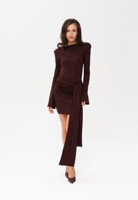 Burgundowa sukienka z długim rękawem o dopasowanym kroju, z marszczonym detalem w talii i wydłużonym drapowanym akcentem. W połączeniu z dopasowanymi szpilkami.