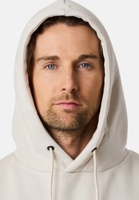 Hoodie beige chiaro con un ampio cappuccio, lacci con punte nere e una texture morbida. Design semplice senza motivi o accenti.