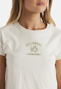 Cremefarvet bomulds t-shirt med rund halsudskæring har et grønt hibiscus blomstermønster og bølgemønster trykt nedenfor "BILLABONG" teksten.
