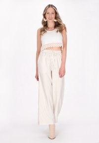 Bílý crop top s krajkovým lemem, spárovaný s pruhovanými, širokými kalhotami v krémových a béžových tónech. Modelka má na sobě béžové kotníkové boty.