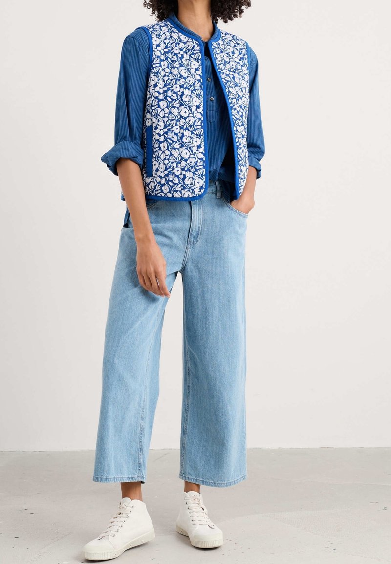 Personne portant une veste matelassée bleue à fleurs sur une chemise bleue à manches longues, un jean large bleu clair et des baskets blanches, debout à l'intérieur.