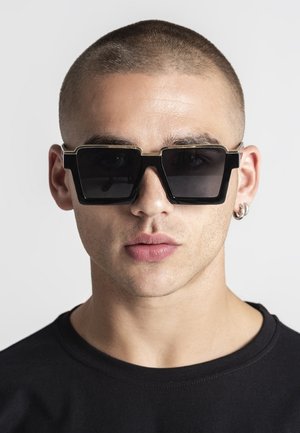 Mladý muž s oholenou hlavou, ktorý má na sebe veľké čierne štvorcové slnečné okuliare, strieborné kruhové náušnice a čierne tričko s okrúhlym výstrihom.