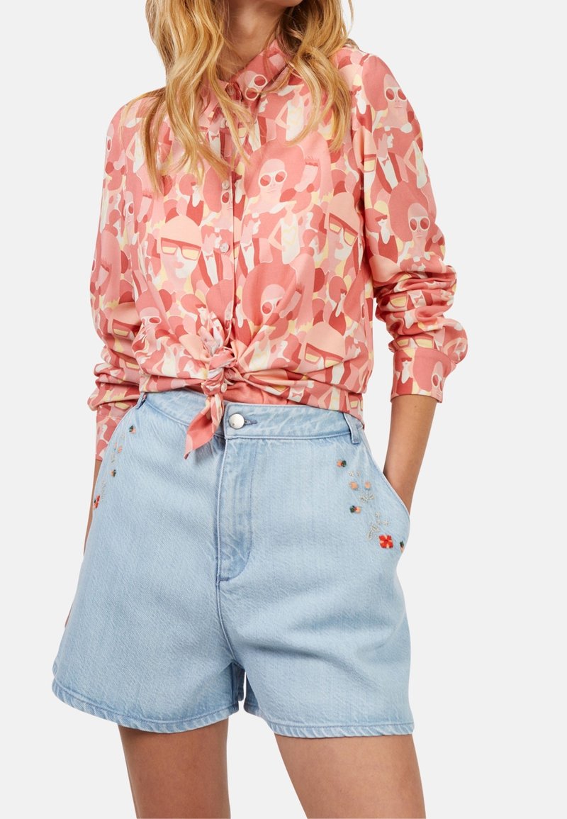 Mujer que lleva una camisa de manga larga abotonada con estampado de caras abstractas rosas y nudo en la parte delantera, combinada con pantalones cortos de mezclilla de tiro alto en azul claro con bordado floral.