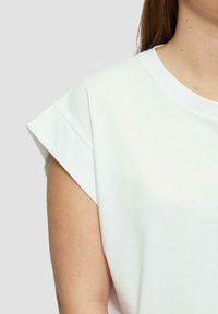 Biała bawełniana koszulka z okrągłym dekoltem, krótkimi, podwiniętymi rękawami i gładką teksturą, skrócona, aby pokazać detal na ramionach i rękawach.