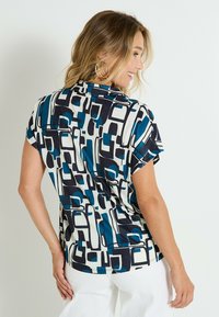Blouse à manches courtes avec un motif géométrique en bleu, marine et blanc. Fabriquée en tissu lisse, avec un col et une coupe décontractée.