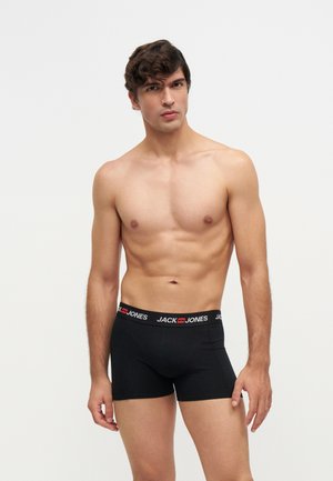 Млад мъж с къдрави тъмни коси, носещ черни боксерки Jack & Jones, стоящ на светъл фон.