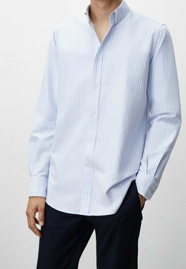 Man in een lichtblauw en wit verticaal gestreept overhemd met zwarte broek, met één hand in de zak tegen een effen achtergrond.