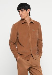 Brown corduroy button-down shirt with a collar, single chest pocket, and green logo detail. Paired with matching brown trousers.