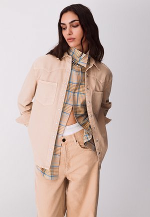 Giovane donna con lunghi capelli scuri, indossa una giacca beige, una camicia a quadri parzialmente fuori dai pantaloni e pantaloni beige, in piedi davanti a uno sfondo chiaro e uniforme.