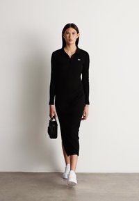 Μαύρο φόρεμα με μακριά μανίκια και πόλο γιακά, πλαϊνές σχισμές και λογότυπο Lacoste. Το μοντέλο κρατάει μια μικρή μαύρη τσάντα και φοράει λευκά αθλητικά παπούτσια.