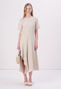 Beige asymmetrisk kjole med korte ermer, teksturert stoff og en myk fall, kombinert med en liten vevd veske og nøytrale hæl sandaler.