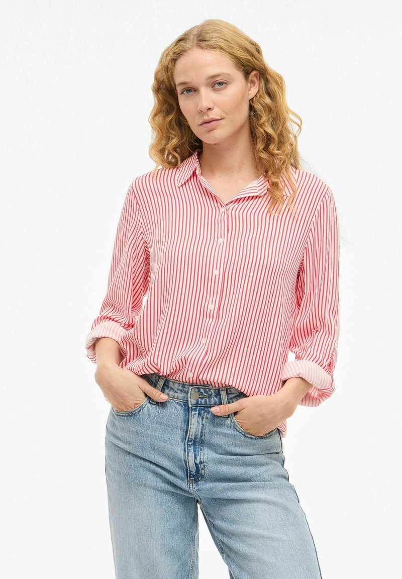 Blouse rayée rouge et blanche avec un devant à boutons, des manches retroussées et une coupe décontractée, associée à un jean bleu taille haute.