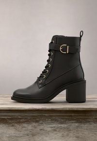 Bottine en cuir noir avec talon bloc, lacets, bride à boucle métallique et languette d’extraction sur surface en bois avec fond uni.