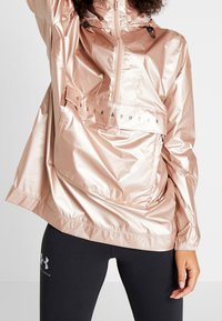 Jasnoróżowa, metaliczna kurtka wiatrówka z półzamkiem, regulowanym kapturem i przednią kieszenią. Posiada pasek z logo na wysokości klatki piersiowej oraz elastyczne mankiety.