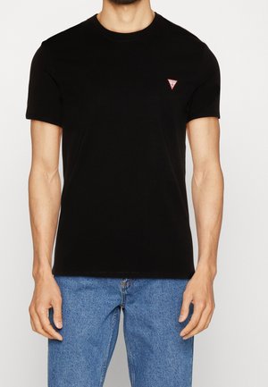 Homme portant un t-shirt noir uni à manches courtes avec un petit logo triangulaire rouge et blanc au niveau de la poitrine gauche, associé à un jean bleu.