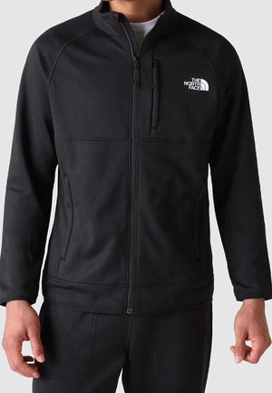 Mężczyzna noszący czarną polarową kurtkę zapinaną na zamek błyskawiczny z logo The North Face na piersi oraz zapinaną kieszenią na piersi, w połączeniu z czarnymi spodniami.