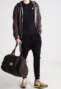Sudadera con cremallera gris oscuro, camiseta negra, pantalones deportivos negros, bolsa de viaje marrón y zapatillas en tonos beige y blanco. Conjunto atlético sencillo y casual.