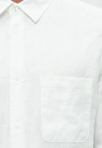 Camisa de linho branca com gola de aba abotoada, botões frontais e bolso no peito com bordas arredondadas. Textura suave com acabamento ligeiramente translúcido.