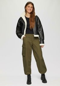 Zwarte leren cropped jas met een witte faux fur kraag, gecombineerd met olijfgroene cargo broek met meerdere zakken en zwarte veterschoenen.