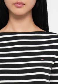 Svart-og-hvitstripet topp med bred utrigning, laget av myk stoff. Har en liten logo på brystet. Jevn tekstur og figursydd design.