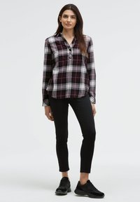 Woman standing wearing a purple and white plaid long-sleeve shirt, black skinny pants, and black sneakers against a plain background.