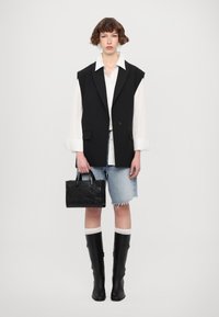 Blazer sans manches noir sur une chemise blanche, associé à un short en jean. Le mannequin porte un petit sac à main noir texturé et des bottes noires jusqu'au genou.
