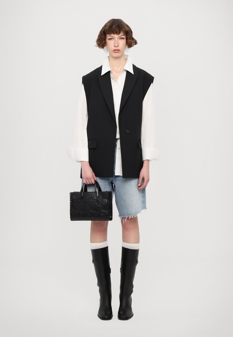 Blazer sans manches noir sur une chemise blanche, associé à un short en jean. Le mannequin porte un petit sac à main noir texturé et des bottes noires jusqu'au genou.