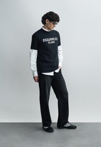 Melnā grafiskā t-kreklā ar uzrakstu “DSQUARED2 MILANO” baltā krāsā, uzvilkts virs baltas garroku krekls. Tumši zili džinsa bikses un melni apavi ar baltām svītrām.