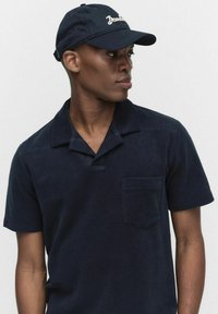 Marineblauwe katoenen polo met korte mouwen, voorzien van een notch-kraag en een borstzak. Gecombineerd met een bijpassende pet met geborduurd logo.