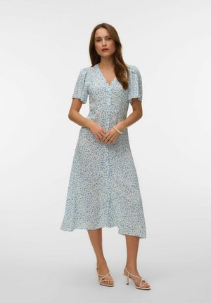 Kobieta w jasnoniebieskiej kwiatowej sukience midi z krótkimi bufkowatymi rękawami i dekoltem w serek, ubrana w beżowe sandały na obcasie, stojąca na jednolitym tle.