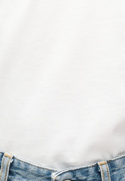 Κοντινό πλάνο ενός ατόμου που φοράει ένα απλό λευκό πουκάμισο, μπαλωμένο μέσα σε μπλε τζιν με εμφανή ραφές και κουμπί.