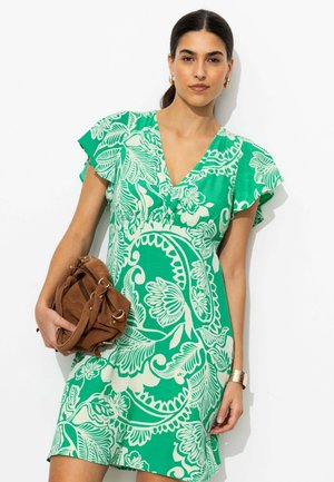 Femme portant une robe courte à manches courtes à fleurs vertes et blanches, tenant un sac à main marron, avec les cheveux attachés et des boucles d’oreilles créoles dorées.