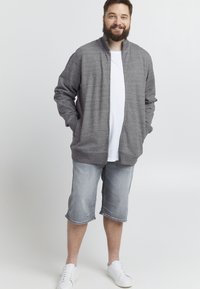 Giacca grigia con zip e colletto alto, abbinata a una maglietta bianca e pantaloni corti grigi chiari fino al ginocchio. Indossata con sneakers bianche.
