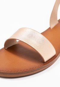 Gros plan d'une sandale en cuir marron avec une large bride beige brillante à l'avant et des coutures visibles le long du bord de la semelle.