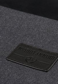 Harmaa kangas, jossa on hieno rakenne, ja siinä on mustasta nahasta valmistettu etiketti, jossa on kohokuvioituna "JOHNNY URBAN" sekä pyöreä logo.