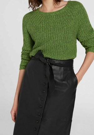 Femme portant un pull en maille vert rentré dans une jupe noire à boutons taille haute avec une ceinture nouée, main dans la poche.