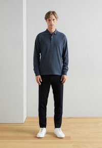 Polo cu mâneci lungi de culoare navy, cu guler, asociat cu pantaloni negri slim și adidași albi, prezentat pe un fundal simplu.