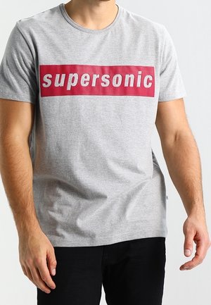 Homme portant un t-shirt gris clair avec une inscription rectangulaire rouge « supersonic » sur la poitrine, accompagné d'un pantalon noir.