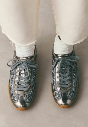 Person wearing white socks and metallic silver sneakers with sequin details and gray laces, standing on a light floor.
