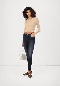 Beige ribbed crop top, dark skinny jeans, leopard print flats, and a white handbag. Model stands in front of a plain background.