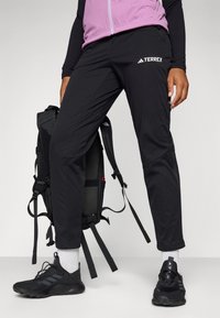 Pantalons Terrex noirs avec poches zippées, associés à des chaussures de sport noires et un sac à dos noir. Tissu texturé avec logos sur le côté.
