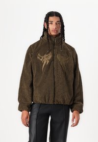 Olivgrön fleecjacka med dragkedja, som har två abstrakta beige mönster på bröstet och elastiska muddar på ärmarna.