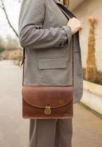 Bruine leren schoudertas met een klep, voorzien van een klittenbandsluiting, gedragen naast een geruite blazer. Gladde textuur en eenvoudig ontwerp.