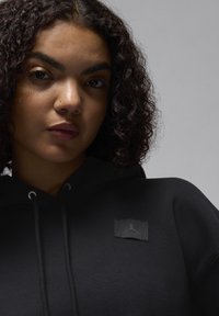 Hoodie preto feito de tecido macio, apresentando um patch de logo texturizado e detalhe de cordão. Acabamento suave com um corte descontraído e bolso canguru.